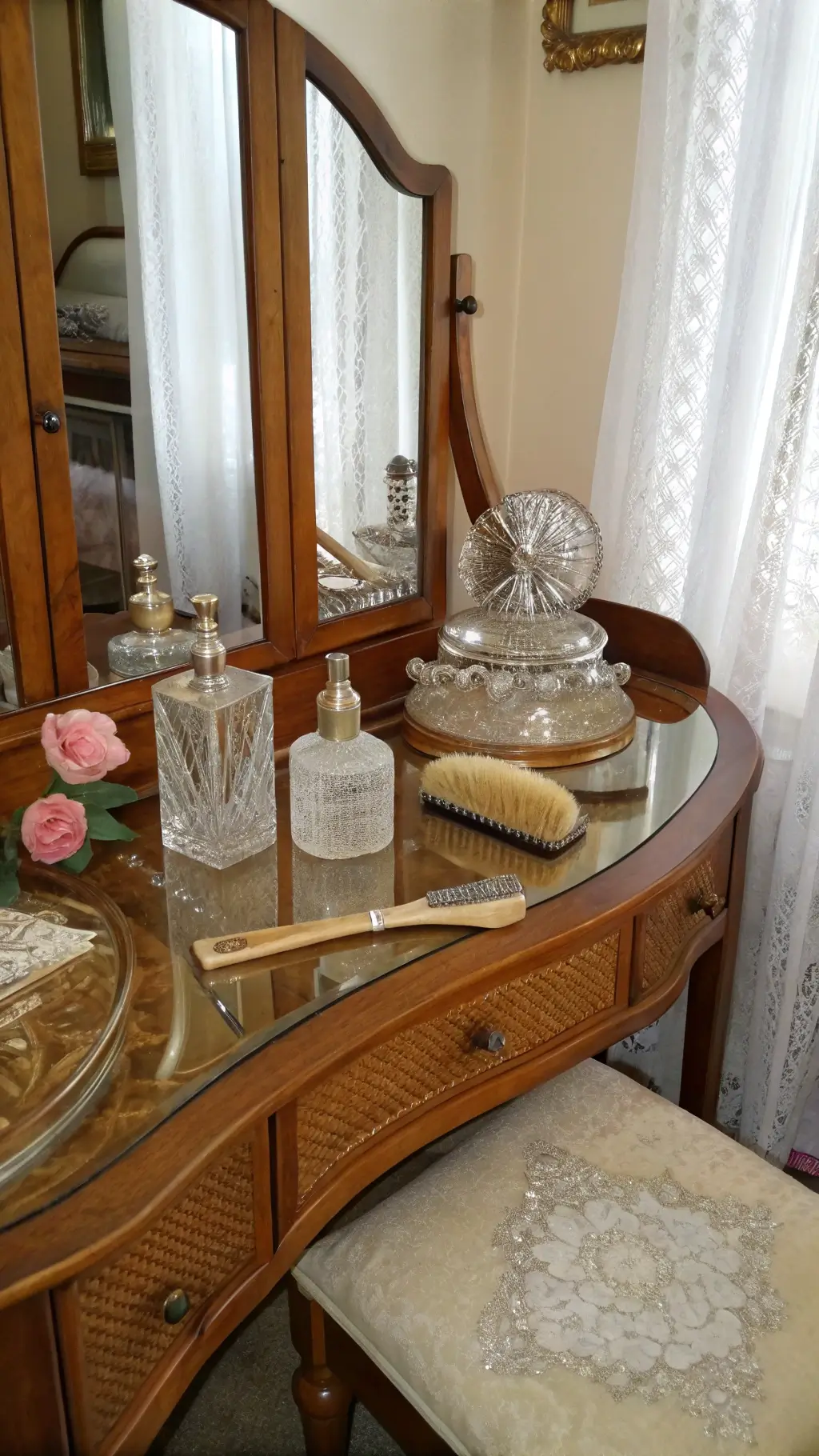 Vintage vanity table in bird's eye maple with a three-panel beveled mirror, silver-backed brushes, cut crystal perfume bottles, and an ivory comb. A rose silk chair sits nearby, with lace curtains softly filtering morning light.