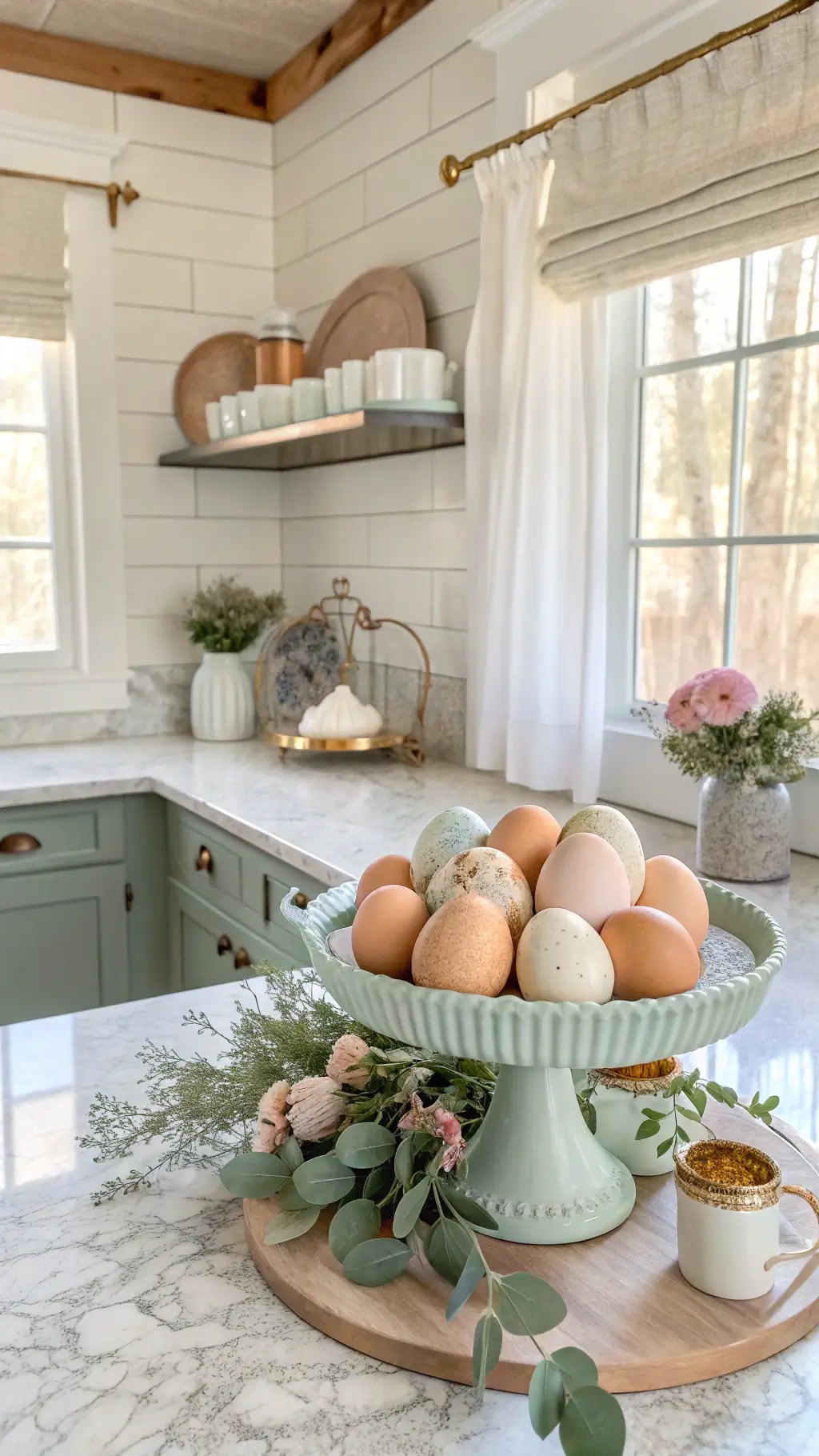 Bright farmhouse kitchen with shiplap walls, marble countertops, and morning light streaming through café curtains. A vintage enamel cake stand holds artisanal speckled eggs in soft sage, cream, and dusty blue. Fresh eucalyptus garland weaves through vintage teacups filled with pale pink ranunculus. Warm copper accents complement the layered textures in a clean, airy composition.