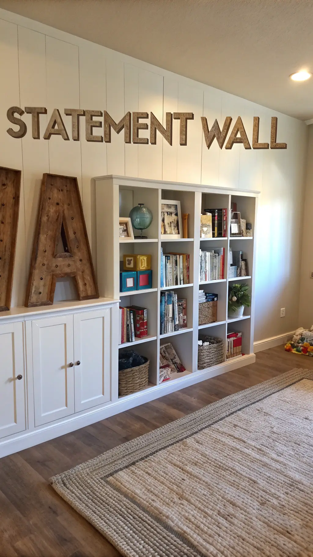 Feature wall with oversized wooden letters in muted metallics illuminated by late afternoon sun. A floor-to-ceiling white bookshelf displays children's books and decorative accessories. A natural jute rug adds texture below.