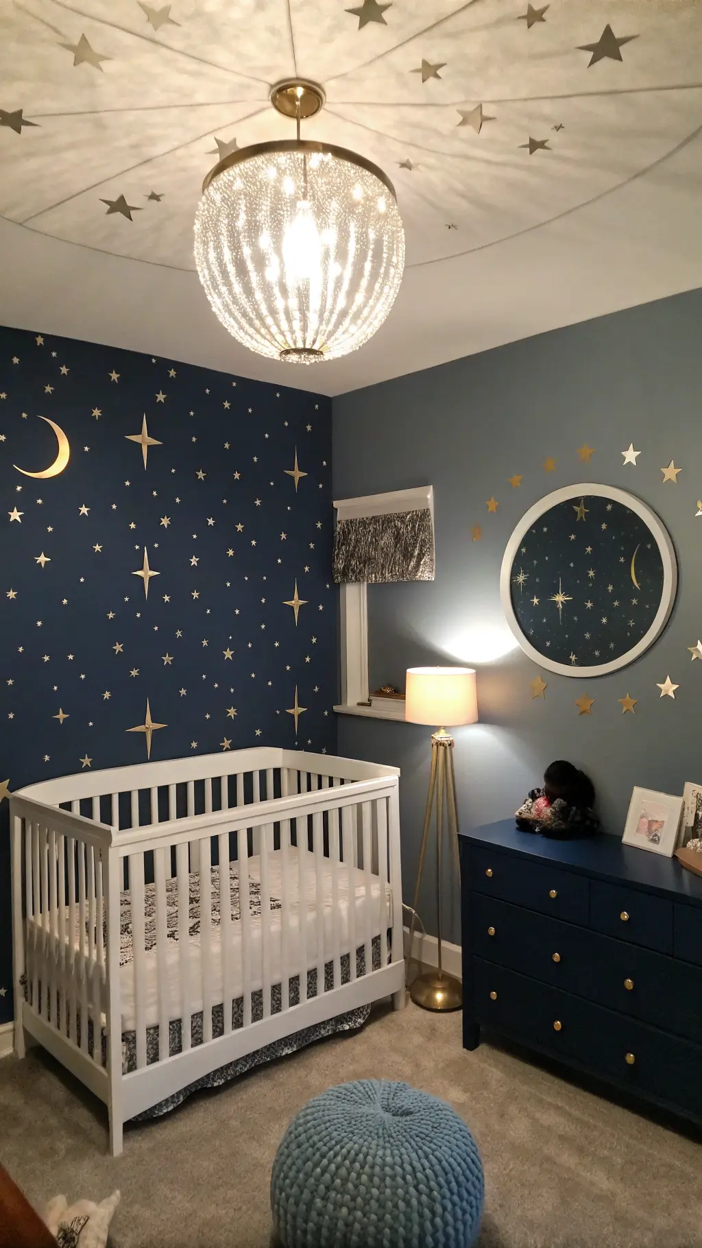 Nursery with navy blue ceiling adorned with metallic silver stars, pearl gray walls with golden moon phase decals, and a white spindle crib beneath a crystal pendant light. A midnight blue velvet glider sits under a round mirror reflecting soft lamplight, creating a magical, celestial ambiance.