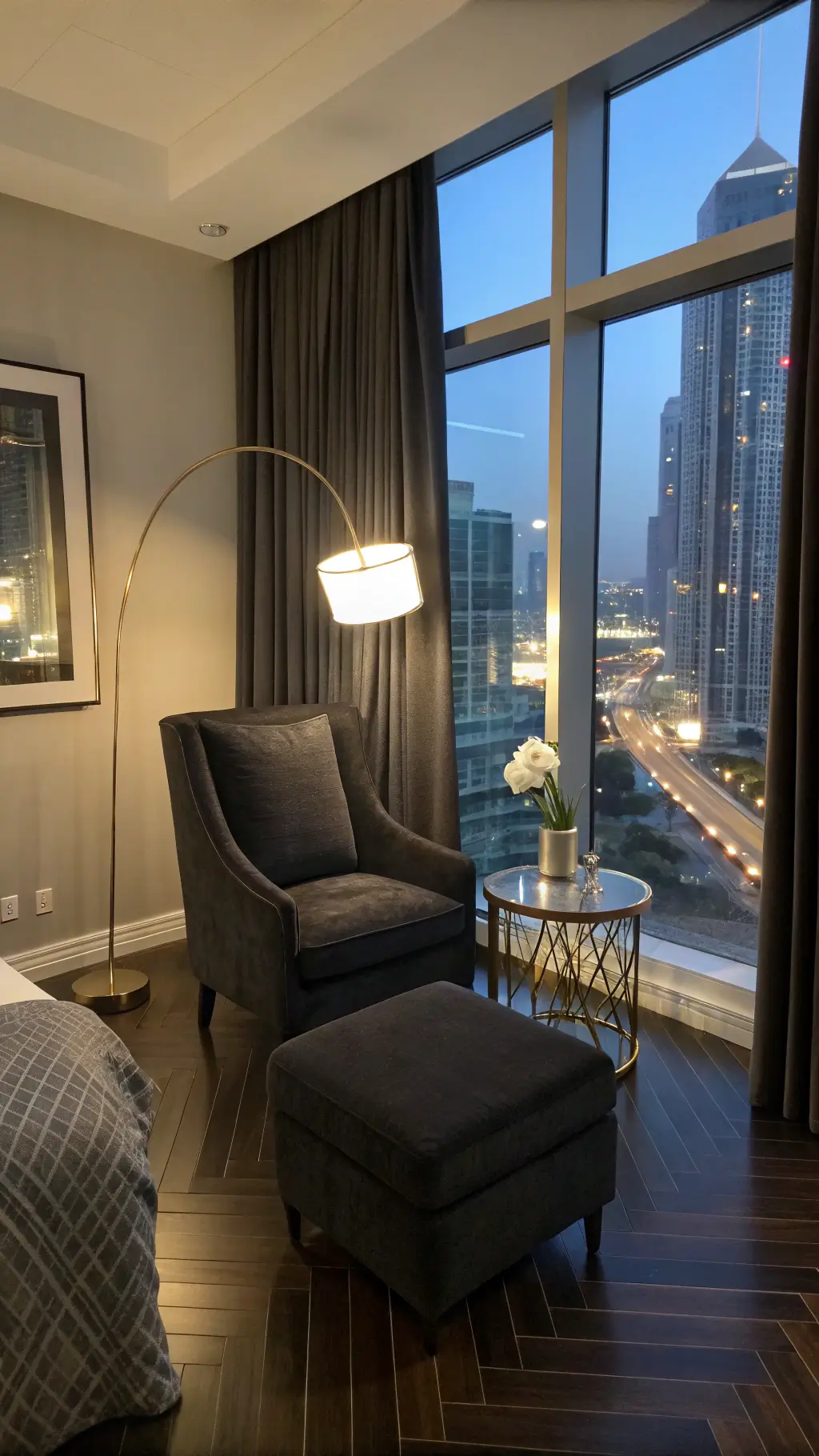 Master bedroom sitting area at twilight with oversized black linen armchair and matching ottoman near a floor-to-ceiling window. Partially drawn blackout curtains reveal city lights. A polished chrome floor lamp casts geometric shadows. A smoky glass side table holds a white orchid. Dark herringbone wood floors contrast with mixed blue hour natural light and warm artificial lighting, creating an urban sophisticated atmosphere.