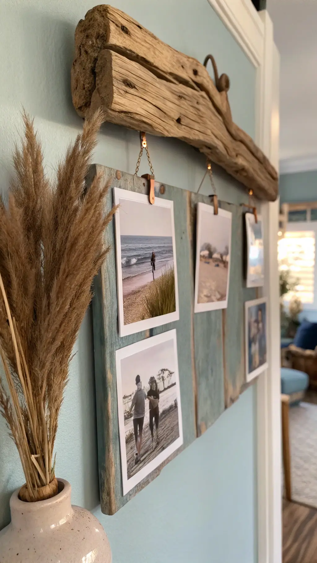Handcrafted driftwood photo display on pale blue-grey wall, with vintage black and white beach photos clipped by copper fixtures. Warm late afternoon light emphasizes the wood’s texture. Below, dried pampas grass rests in a handmade ceramic vase. Soft-focus background hints at a larger room.