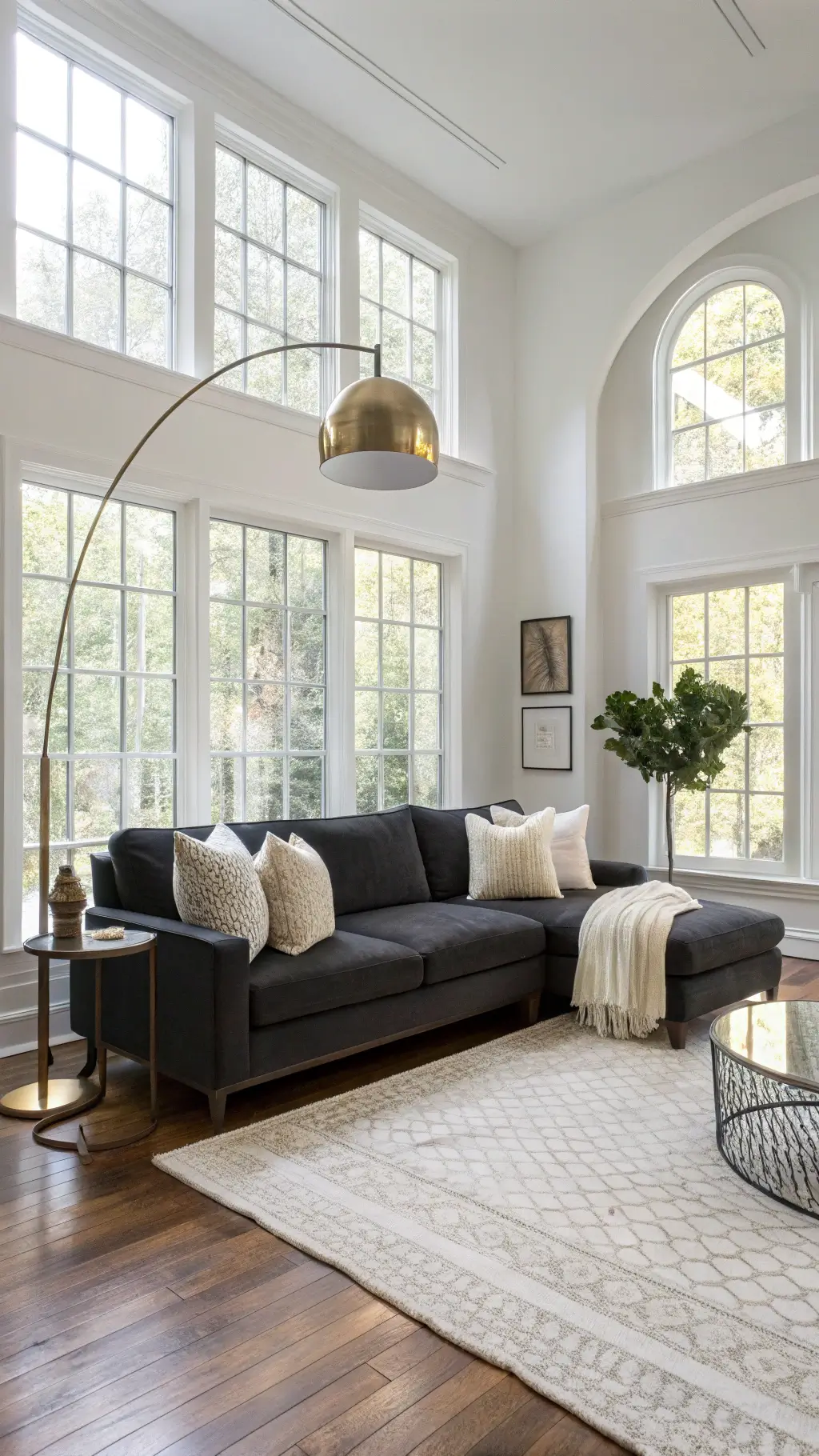 A spacious 20x24ft living room with 10ft ceilings, flooded with golden hour light from large west-facing windows. A dark charcoal L-shaped sofa with cream, white, and beige textured pillows sits on layered cream rugs over walnut hardwood floors. A brass floor lamp arches overhead, while a chunky ivory knit throw and mixed metallic accents add warmth. A large fiddle leaf fig catches the sunlight, enhancing the airy feel.