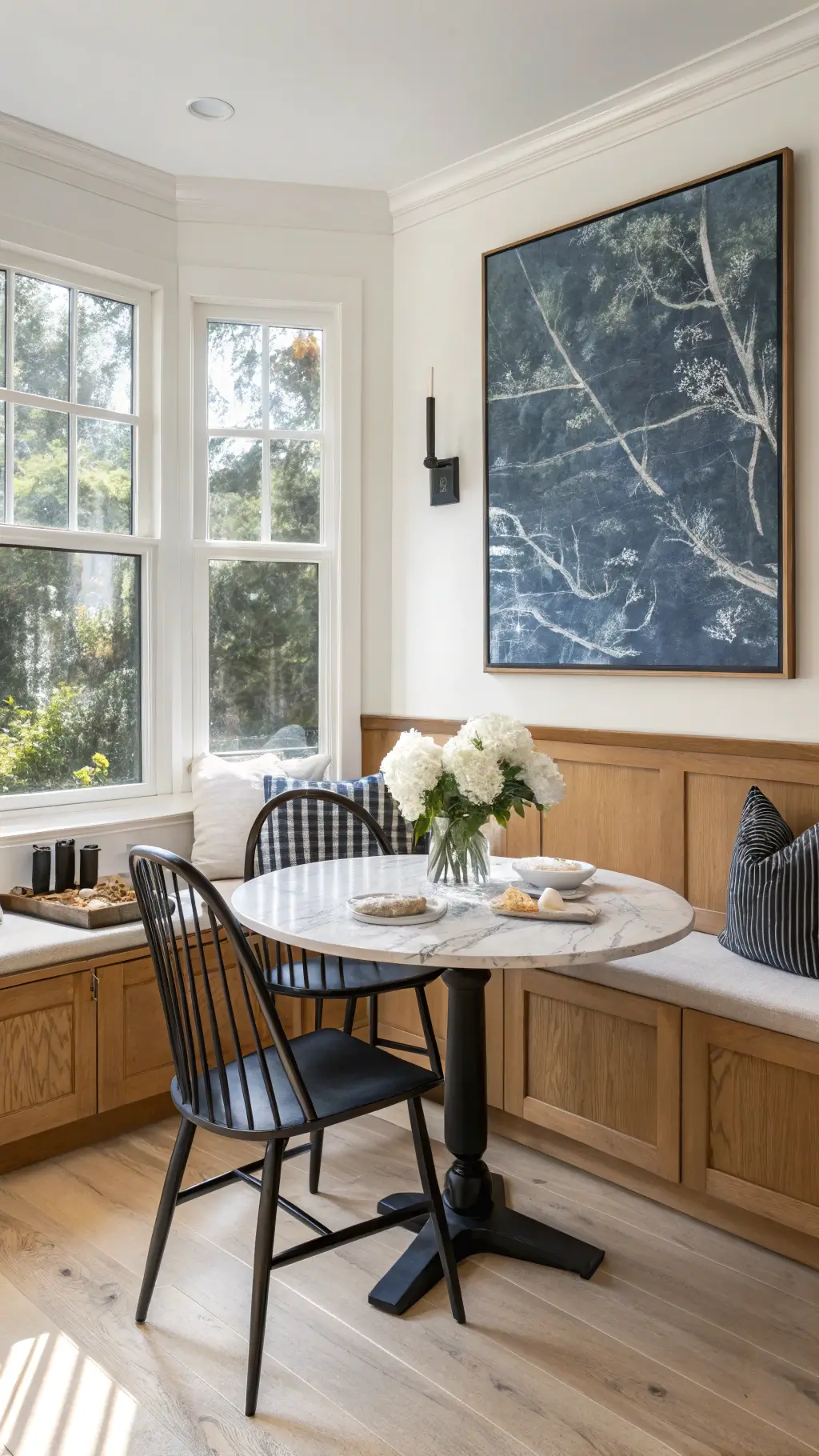 Modern breakfast nook with black Windsor chairs, oak bench seating, white marble table, and large abstract art, bathed in late morning natural light.