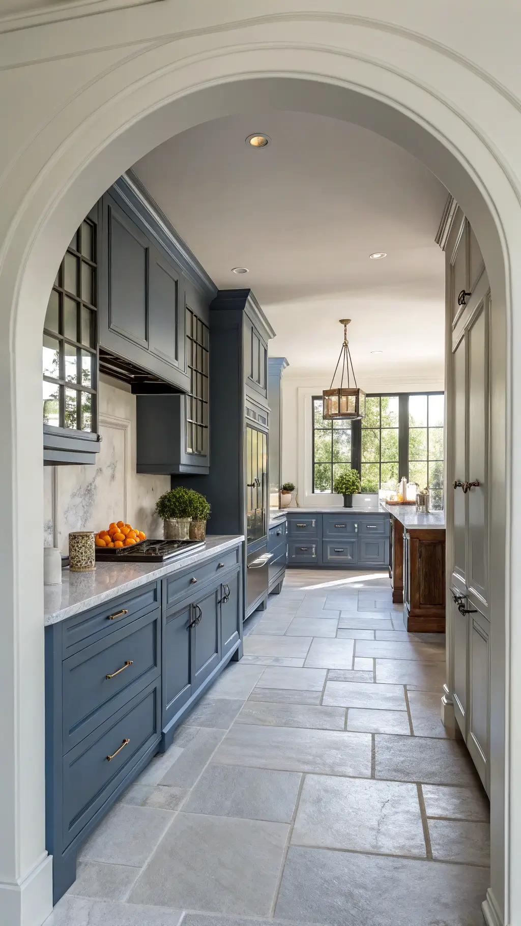 Transitional kitchen with slate blue cabinets, gray limestone floors, and a white marble backsplash; features a quartz waterfall island accented with modern ceramics and fresh citrus, viewed straight-on in soft natural light.