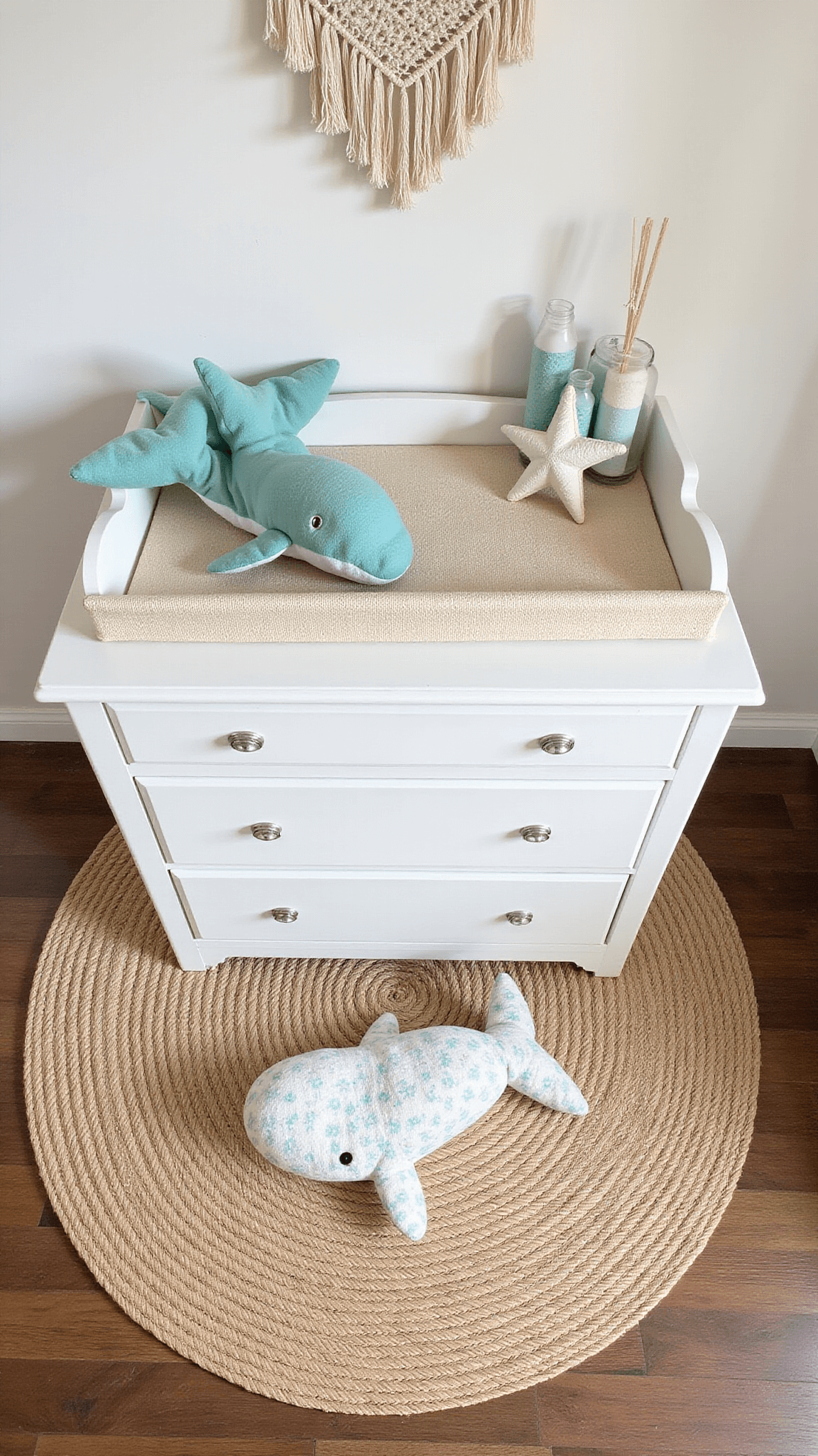 Overhead view of coastal-themed baby changing station with woven pad on white dresser, sea glass-colored essentials, jute rug, stuffed whale, starfish pillow, and macramé wall hanging in soft morning light.
