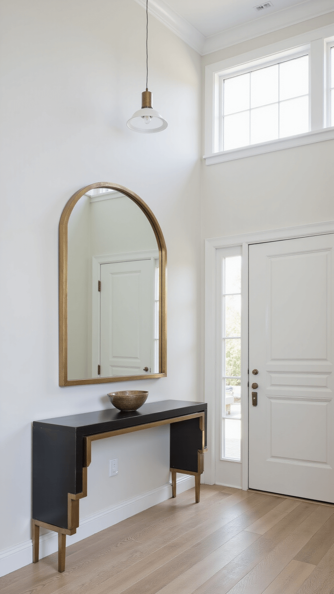 Minimalist 6x8ft entryway with brass-framed mirror, smoked glass console, ceramic vessel, and modern pendant light under 9ft ceiling with transom window.