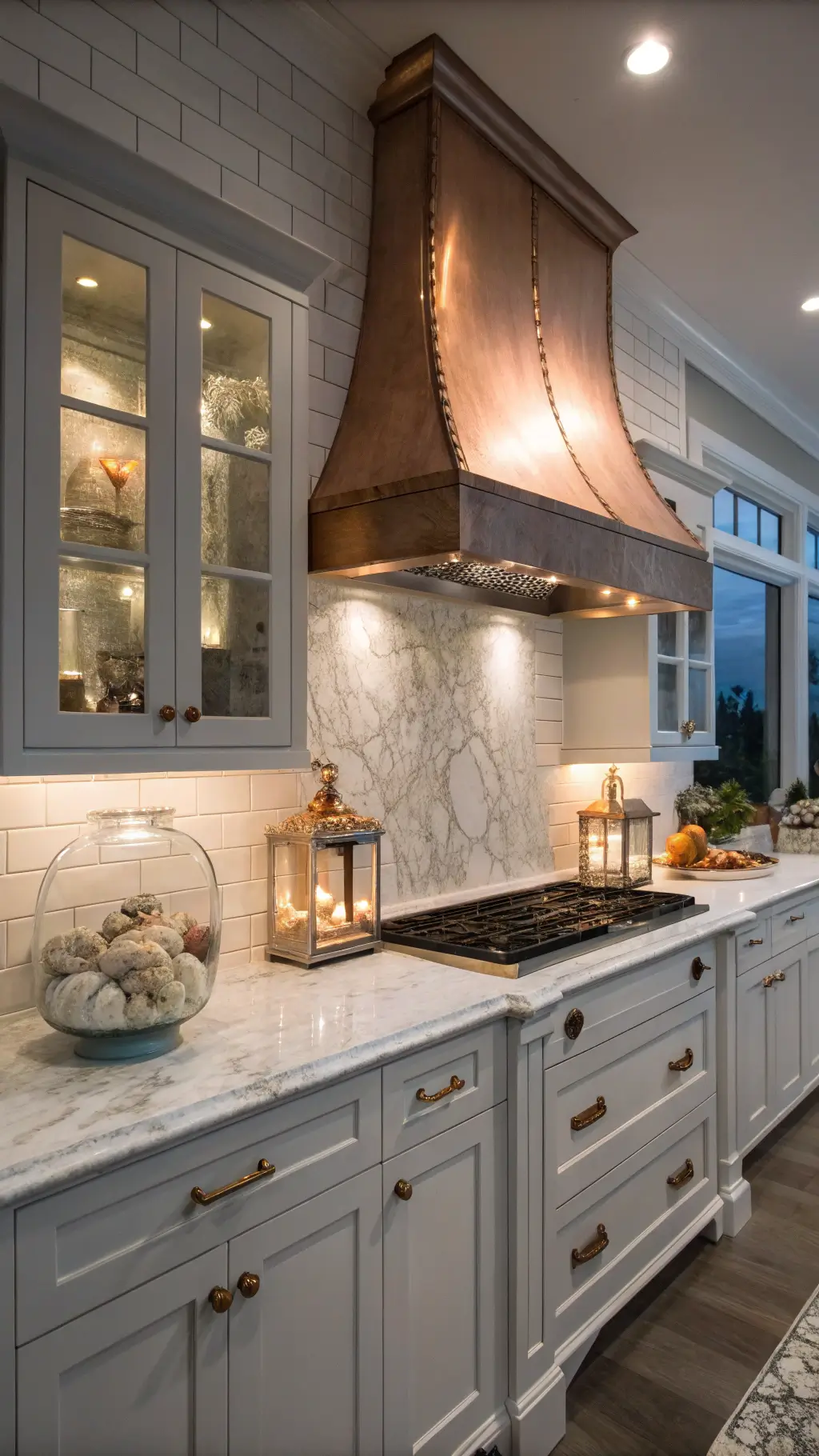 Low-angle view of a twilight coastal kitchen with copper hood, gray cabinets, white subway tile, Carrara marble counters, and lantern sconces highlighting shell and coral displays.