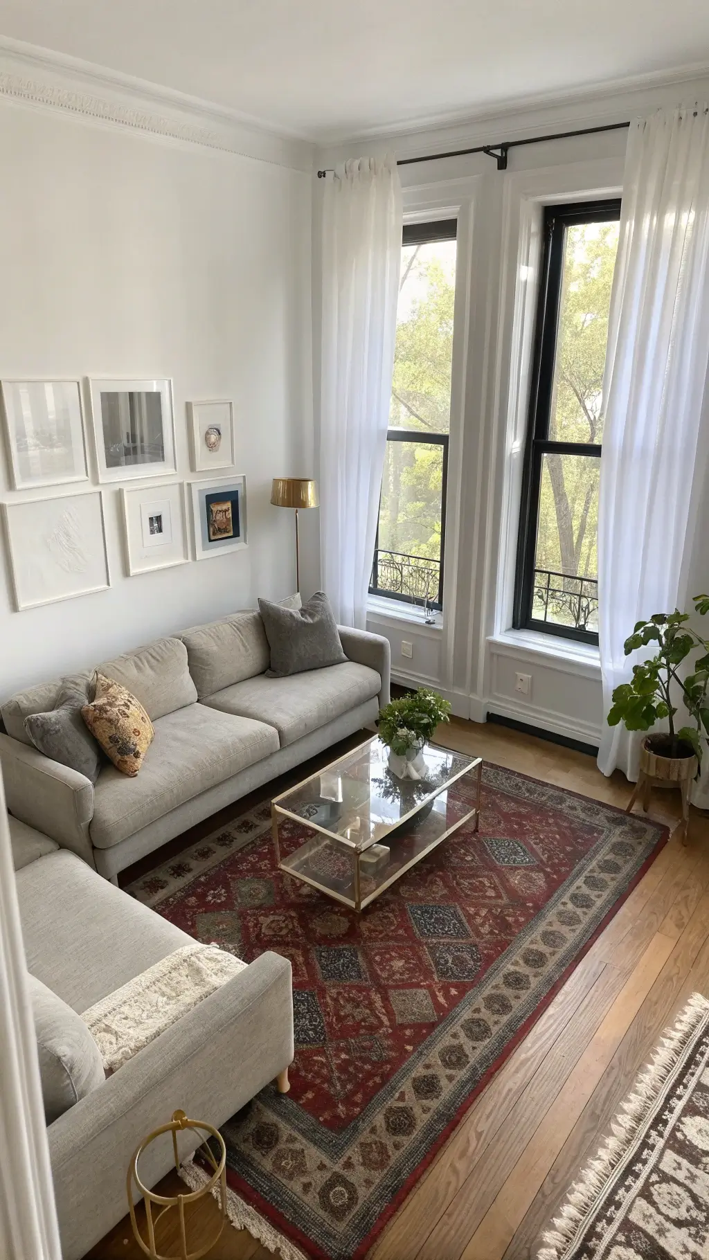 Aerial view of a sunlit 10x12' corner living room with gray linen sofa, lucite nesting tables, white walls contrasting with black-framed windows, brass sculptures, vintage Persian rug, earthenware collection, and mirrors augmenting space.