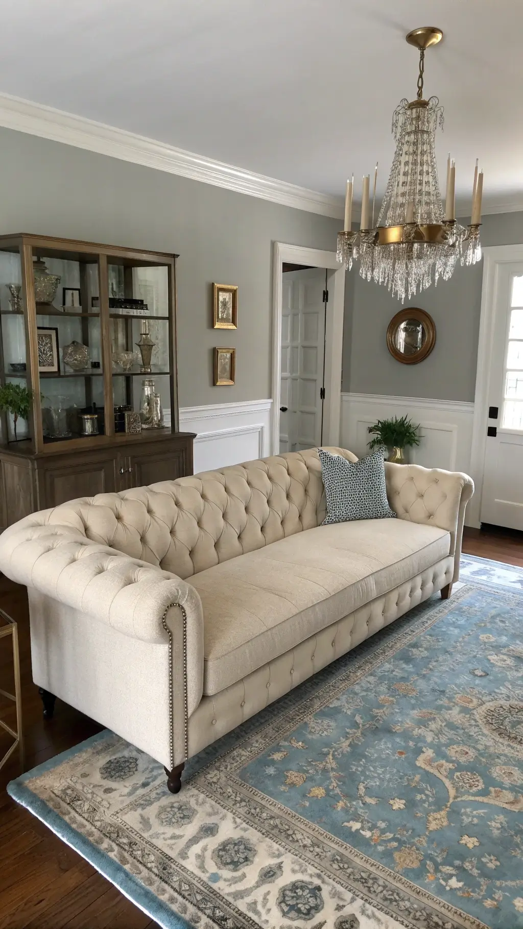 Elevated wide-angle shot of a sophisticated 12x13ft traditional-modern fusion space with an oatmeal linen tufted sofa, brass étagère against pale gray walls, vintage overdyed rug in muted blue-grays, and a crystal chandelier for glamour.